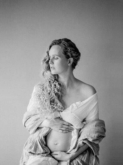 Zwart-wit fine art portret van een zwangere vrouw met gipskruid in het haar en haar handen liefdevol op haar buik.