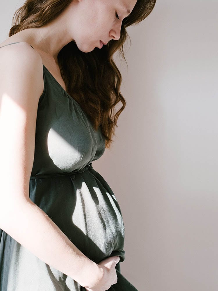 Zwangere vrouw in een groen linnen jurkje die liefdevol naar haar buik kijkt in warm natuurlijk strijklicht.